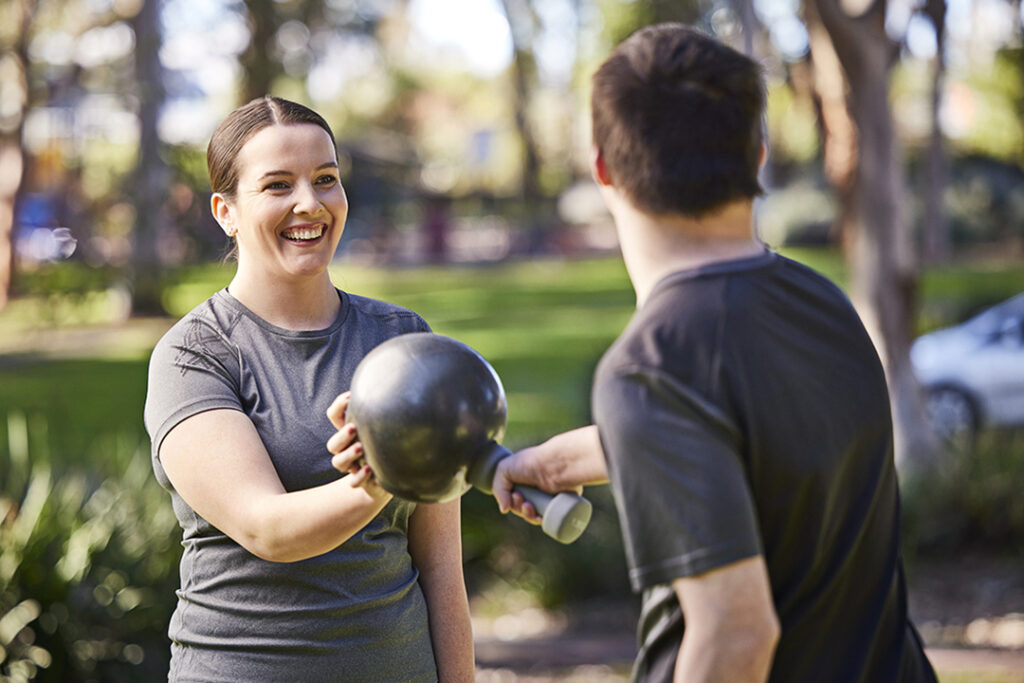 A lady with a medicine ball is smiling while offering personal training to a man wearing a dark shirt