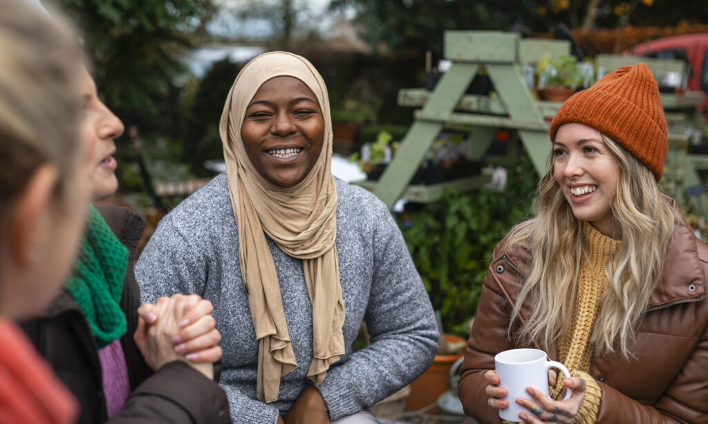 Volunteers sitting outdoors wearing warm casual clothing on a sunny cold winters day. They are resting and having a tea break from working on a community farm, looking after crops and performing other sustainable and environmentally friendly tasks. They are laughing and talking together, drinking hot drinks.