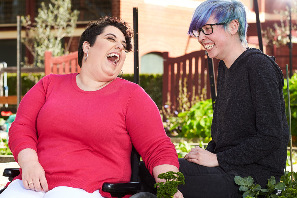 A lady in a wheelchair wearing a pink top laughing with her support worker in a garden