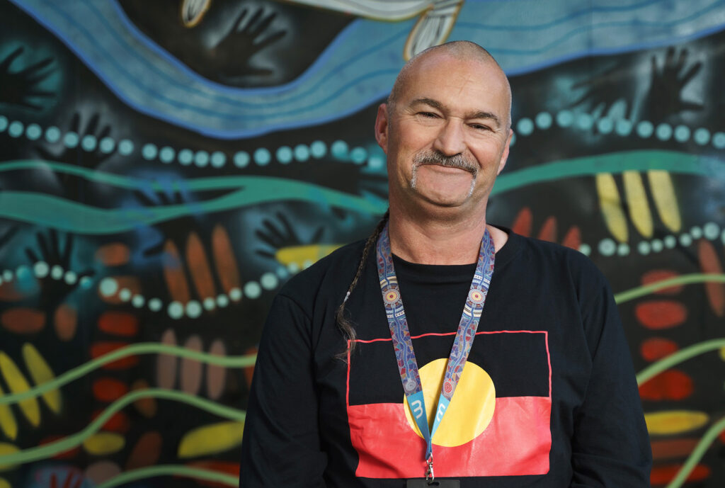 A man in a shirt with an aboriginal flag standing in front of a mural