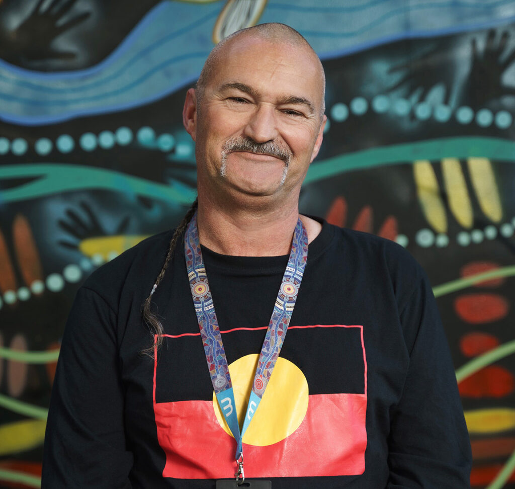 A man in a black shirt with an aboriginal flag standing in front of a mural