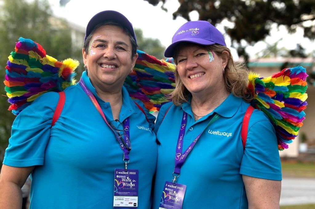 Two women at a pride parade wearing blue Wellways shirts and rainbow feather wings