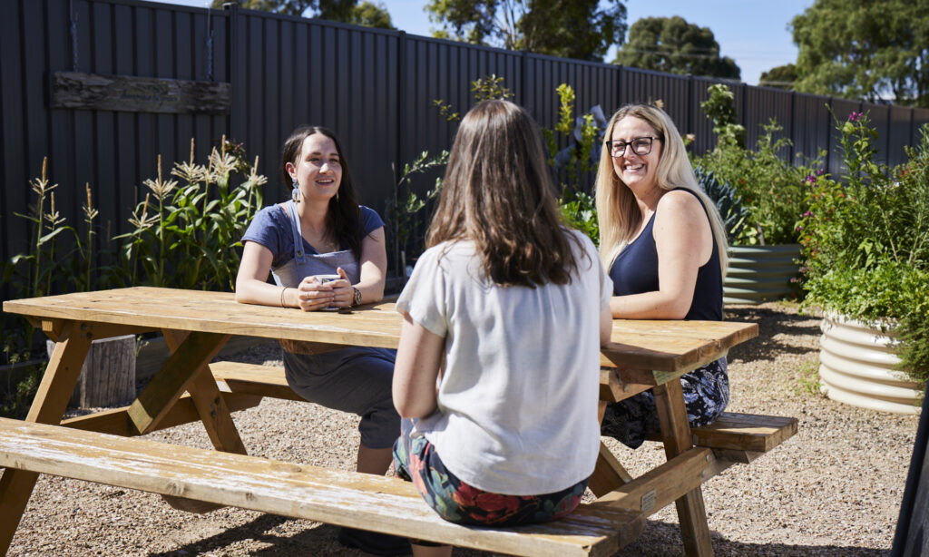 Three people sitting at a garden table in the sunshine talking at Prevention and Recovery Care centre.