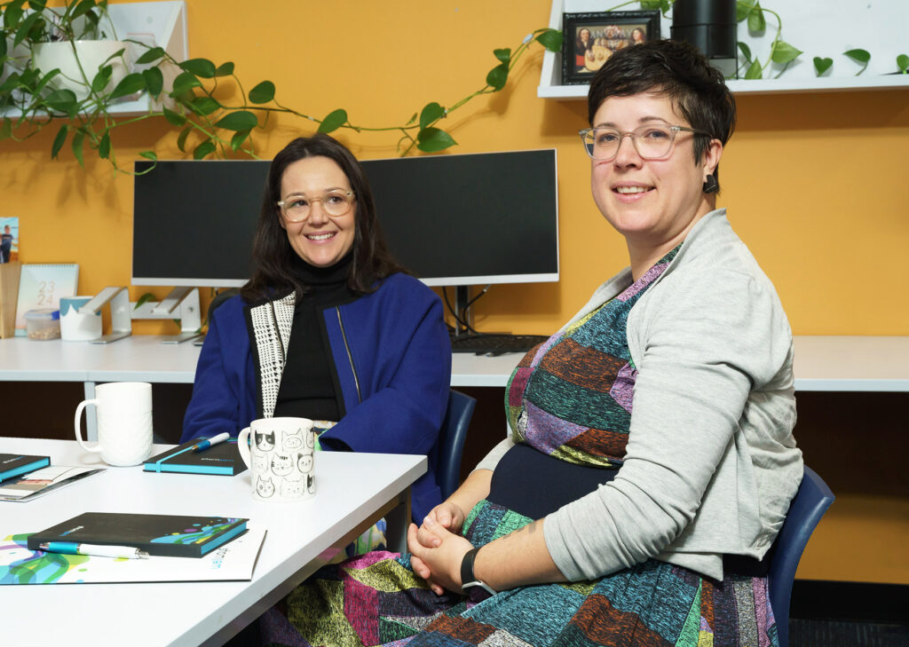 Two staff members sitting at a table smiling with a yellow wall and office plant in the background