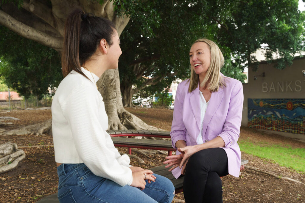 Two people sitting on a park bench under a tree speaking as if in a counselling session.