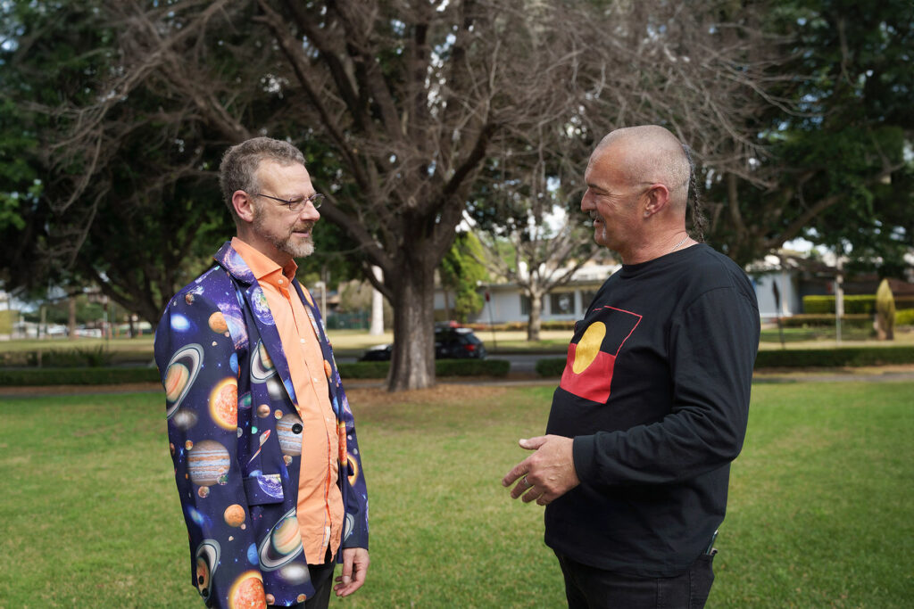 Two people facing each other and talking in a park with a tree in the background.