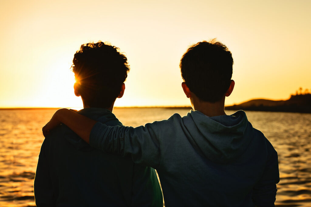 Two boys with their arms around each other watching the sunset over the ocean.