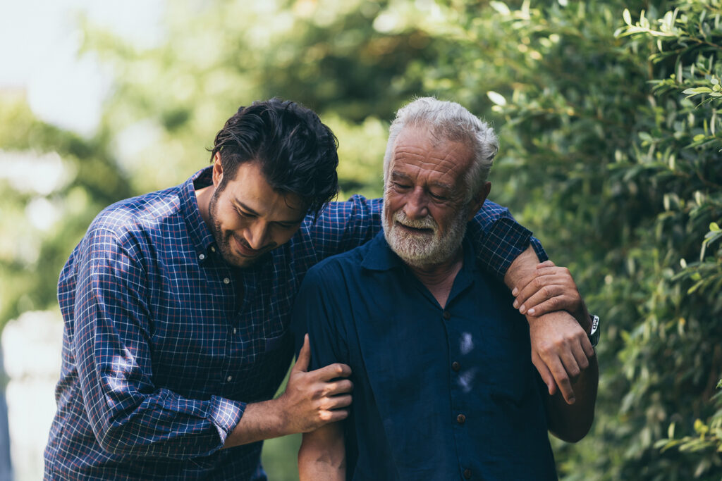 An old man and his son are walking in the park. A man hugs his elderly father. They are happy and smiling