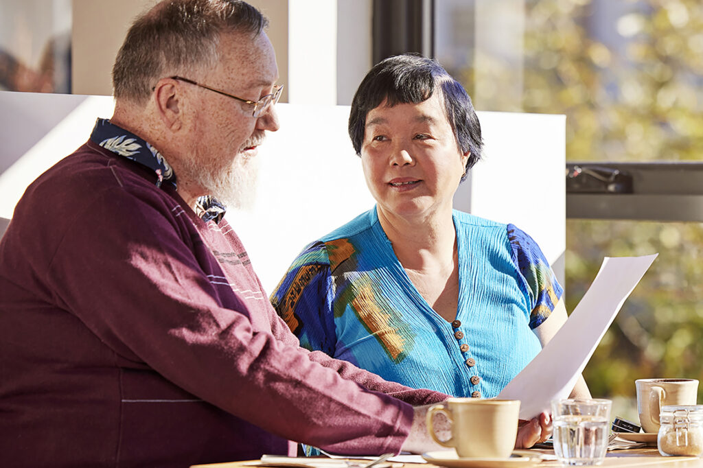 A man reading information to his wife from a piece of paper while sitting at a table with tea cups