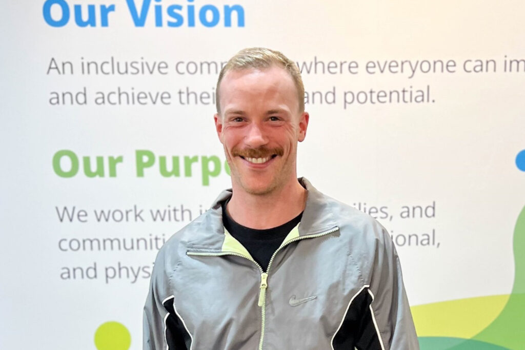 A man with a moustache standing in front of a wall with sign writing