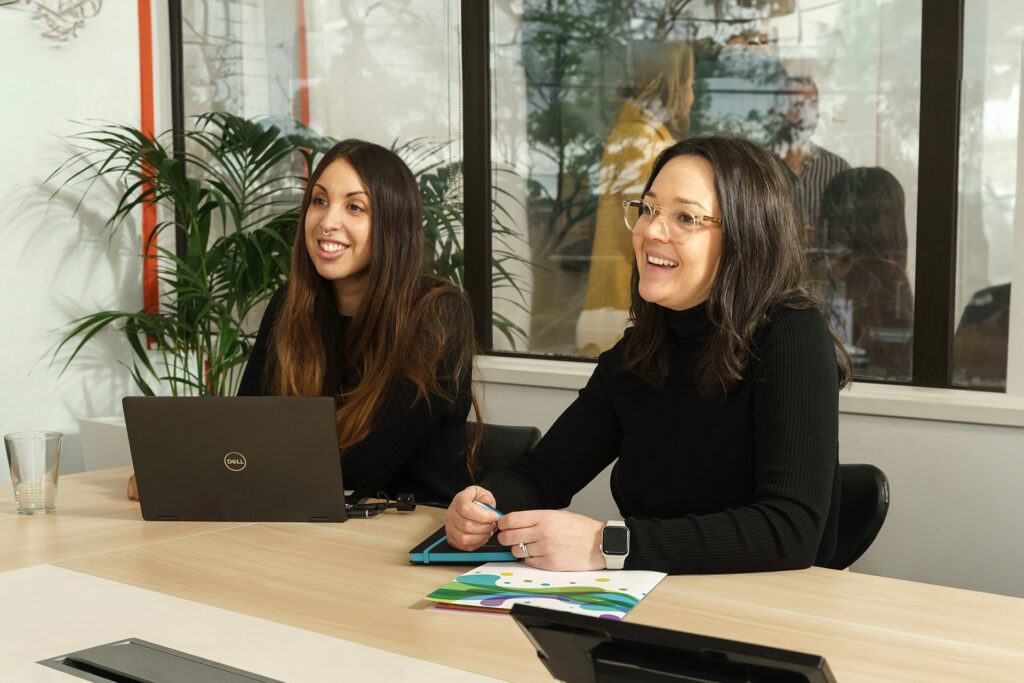 Two ladies sitting at a work desk in a meeting room smiling.