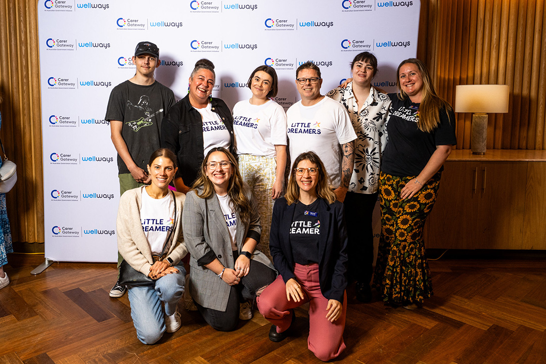 A group of nine people standing and kneeling in front of a media photography wall with Wellways Carer Gateway insignia.