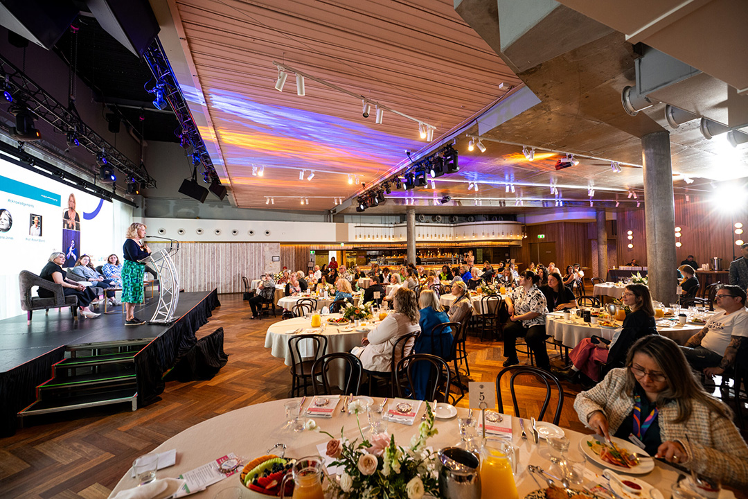 A room full of people sitting at round tables, eating breakfast while watching and listening to people present on stage.