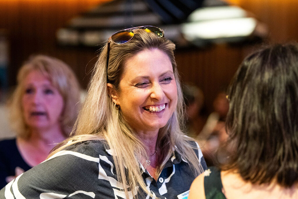 A person with long blonde hair and sunglasses on their head, smiling while chatting with another person.