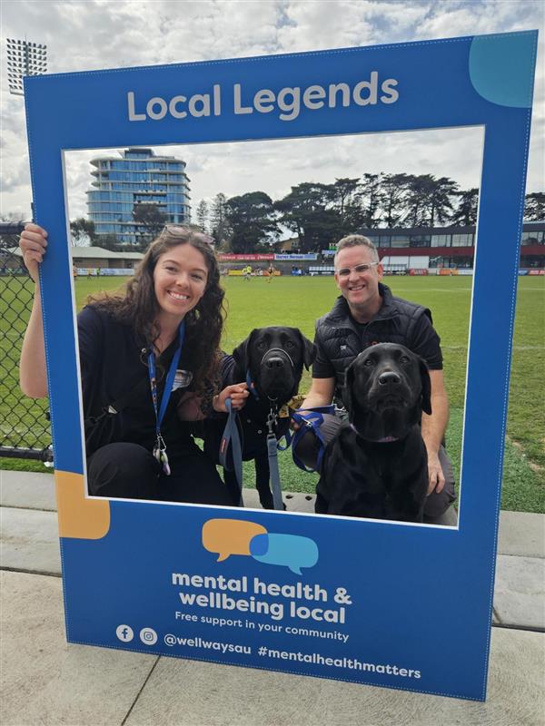 Two people and two dogs posing behind a Blue Frame for a photo