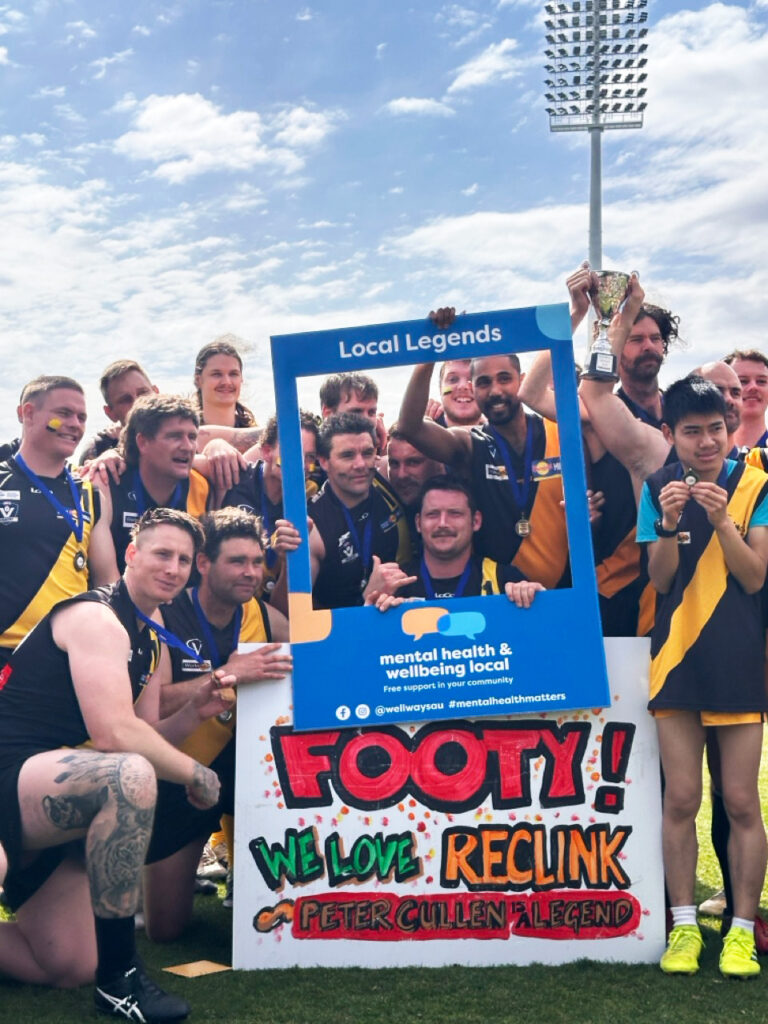 Cardinia tigers football team holding Mental Health and Wellbeing Local Selfie frame in front of a painted sign that says Footy!