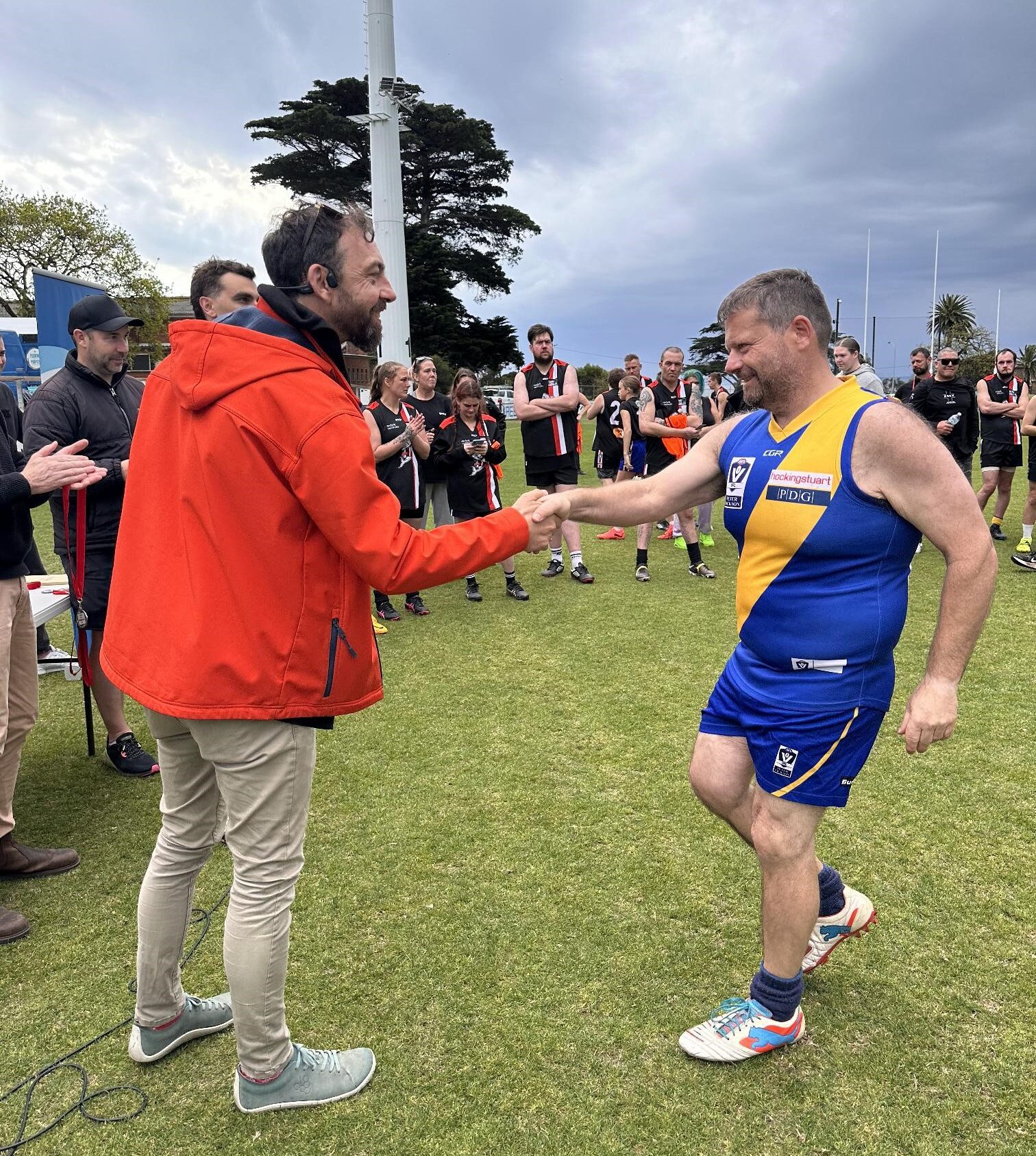 A man in a red jacket shakes the hand of a football player wearing Blue and Yellow jersey.