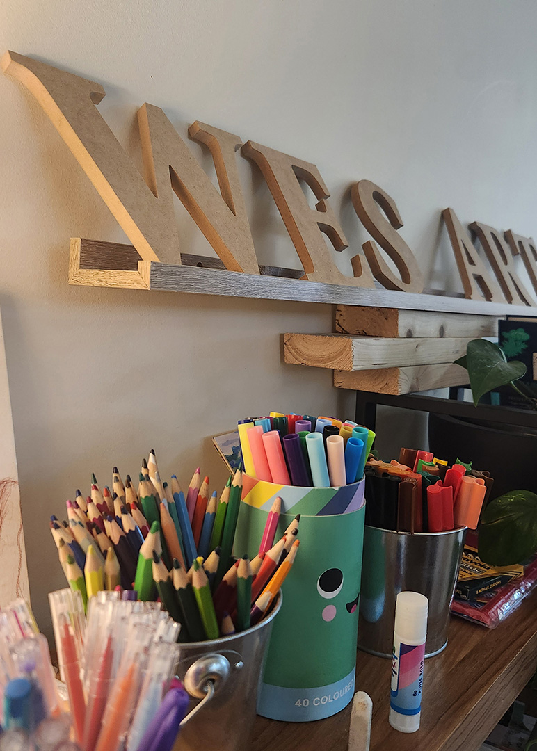 Coloured pencils, markers and other craft supplies on a shelf with the words WES ART in timber behind. 