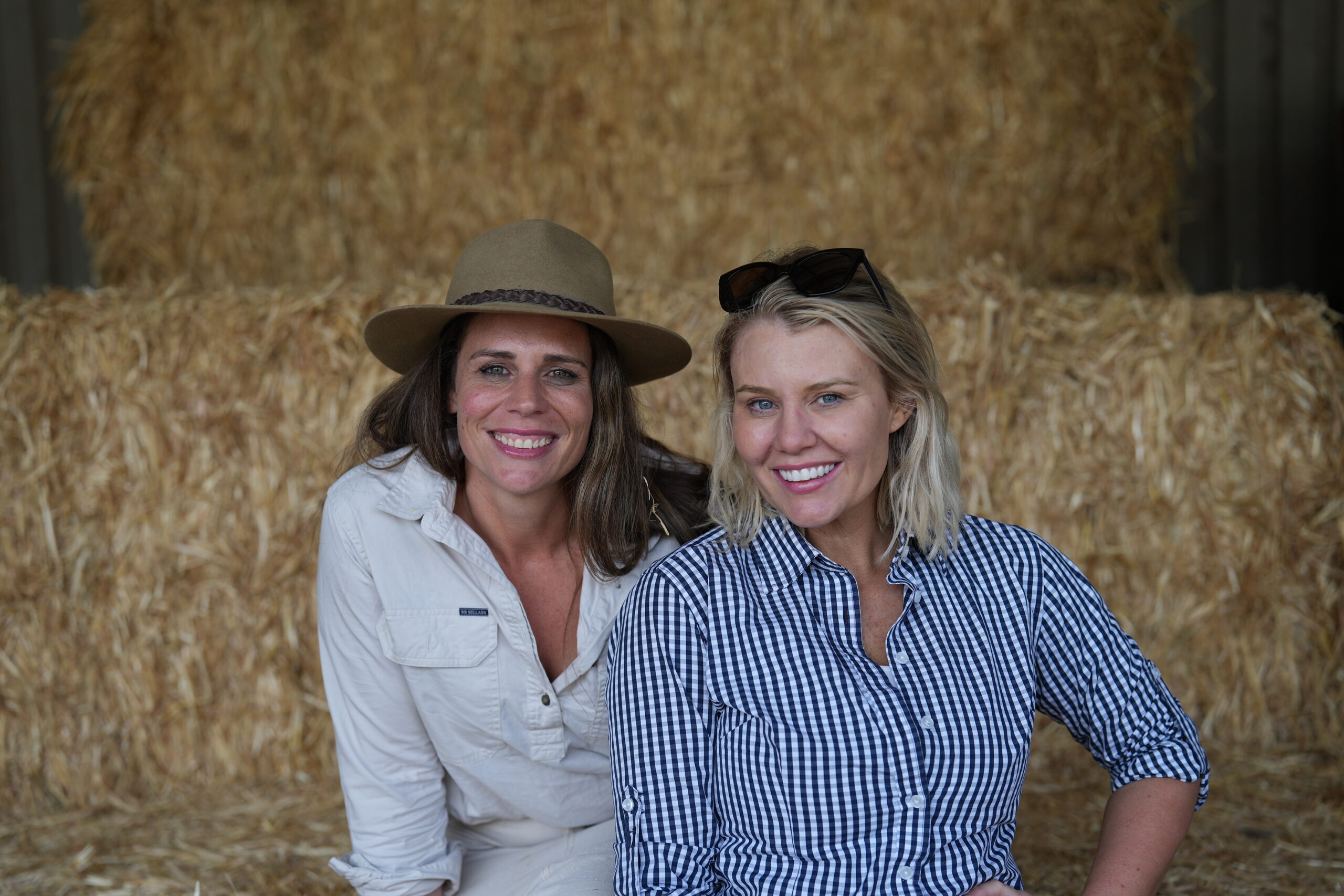 Two people sitting on a hay bale. One is wearing a hat, the other is wearing a blue checked shirt.