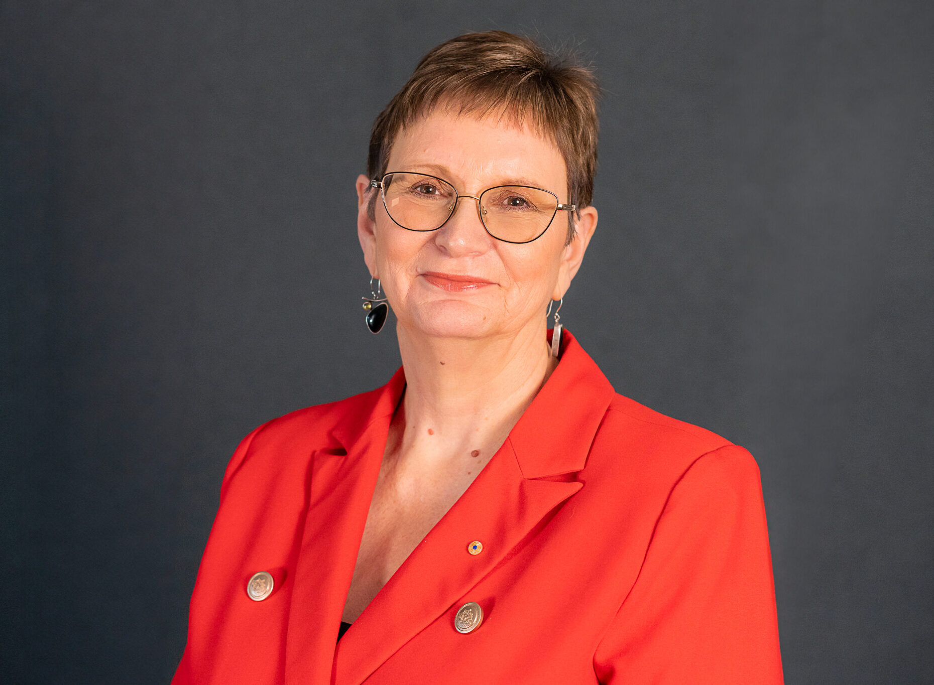 Portrait of Dr. Peggy Brown AO wearing in a red jacket in front of a grey wall. 